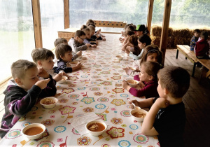 Dzieci z najstarszej grupy siedzą przy długim stole i jedzą zupę pomidorową.